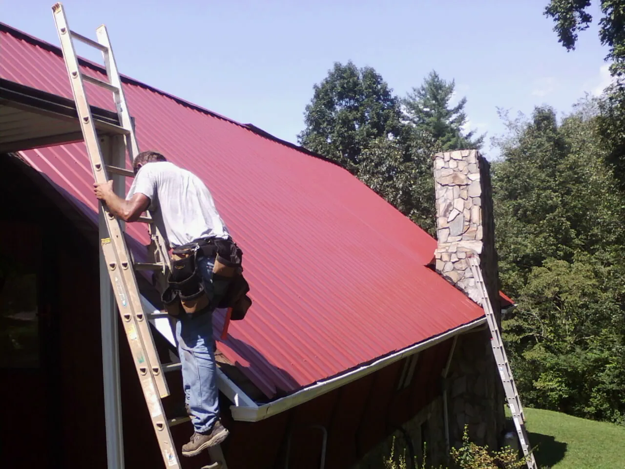 Roofer on a ladder beside a red metal roof and stone chimney — Soffit & Fascia Repair by Roofing Brothers in Sachse
